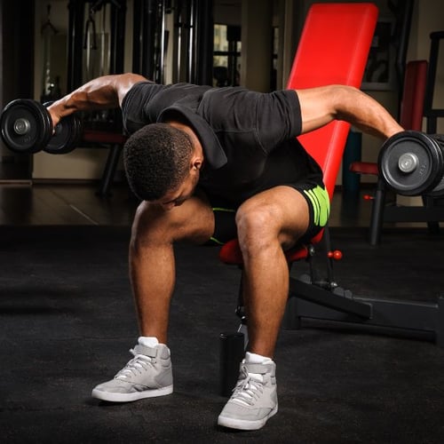 Man seated on weight bench performing bent-over lateral raises with dumbbells