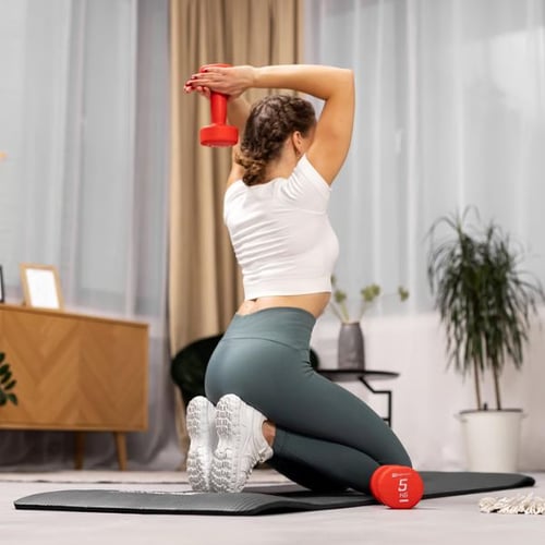 Woman kneeling on mat doing overhead triceps extension with red dumbbell