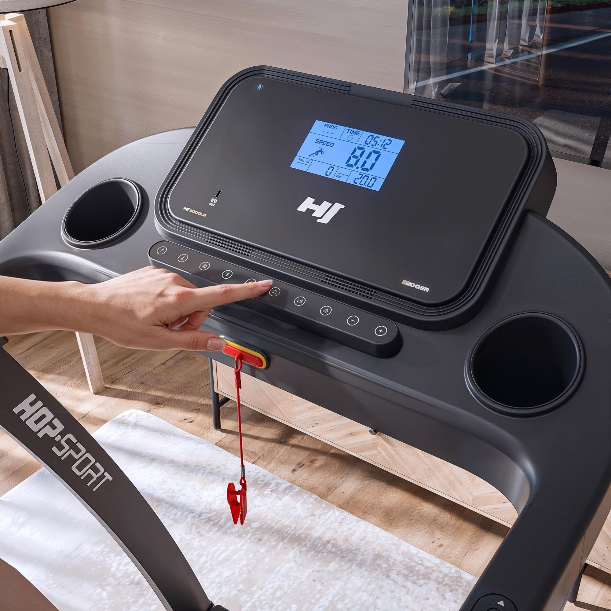 Close-up of a treadmill's electronic console featuring a blue backlit LCD display showing workout statistics like speed and time. A hand is pressing a button on the control bar below the screen, next to two integrated cup holders and a red safety key clip.
