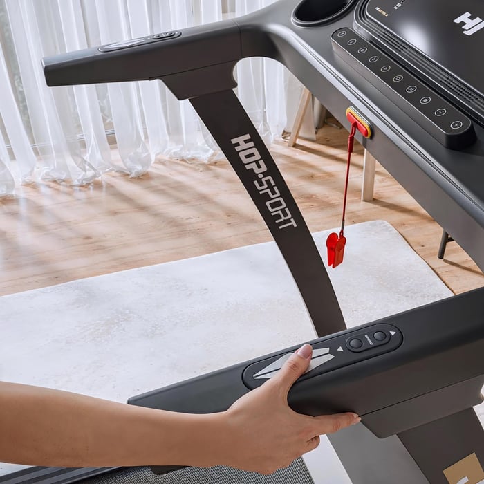 Close-up of a person's hand holding the side handle of a treadmill. The handle features integrated heart rate sensors and thumb-operated speed adjustment buttons for safe and easy control during a workout.