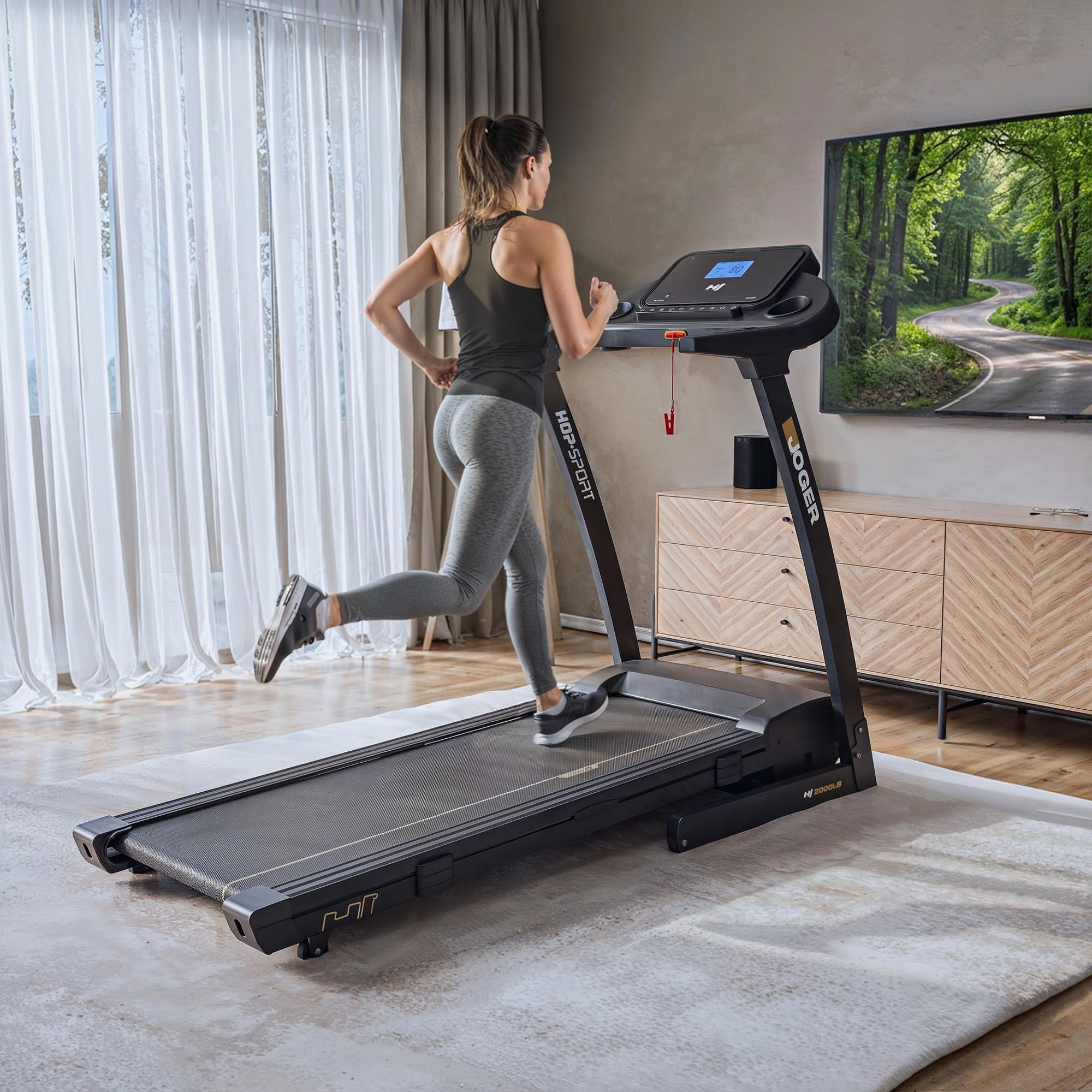 A woman in gym wear lifting the running surface of a foldable treadmill towards the console to save space. This demonstrates the equipment's compact storage functionality within a home interior.